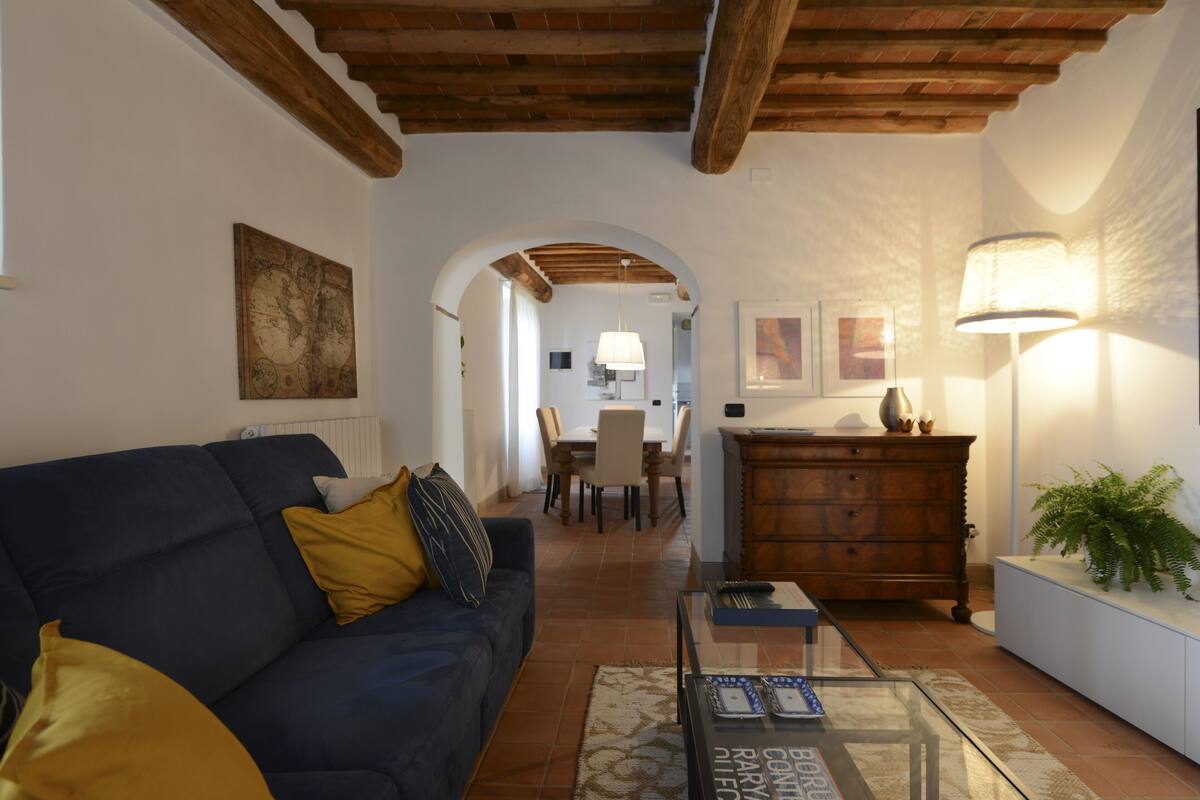 Una sala de estar con un sofá azul oscuro, un aparador de madera, una mesa de centro de vidrio y un comedor al fondo.