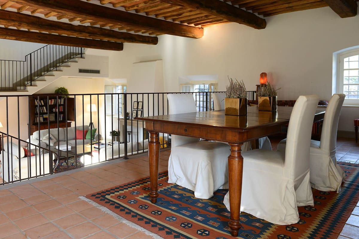 Un comedor con una mesa de madera y sillas blancas tapizadas, un rug con diseño y una escalera que sube a otro nivel.