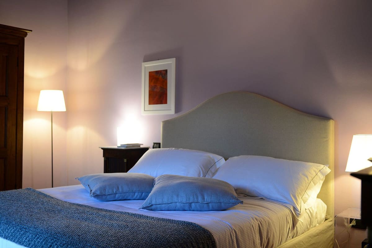 Una cama bien tendida con cabecera gris, dos lámparas de noche, un cuadro enmarcado en la pared y una puerta de madera.