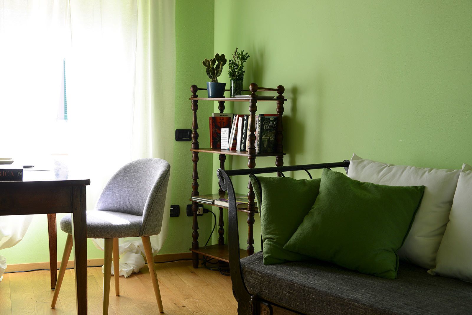 Una habitación con una silla de madera, un sofá verde con cojines, una estantería de madera con libros y plantas, y una ventana con cortinas.