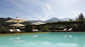 A swimming pool with lounge chairs and umbrellas, surrounded by greenery and mountains in the background. - Guesthouse ai Santinelli (Capannori)
