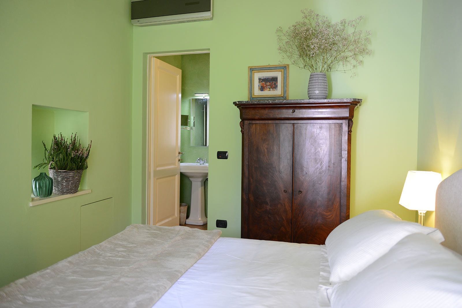 Un dormitorio con cama, un armario de madera, una pared verde, una pequeña estantería con plantas y un baño visible a través de una puerta abierta.