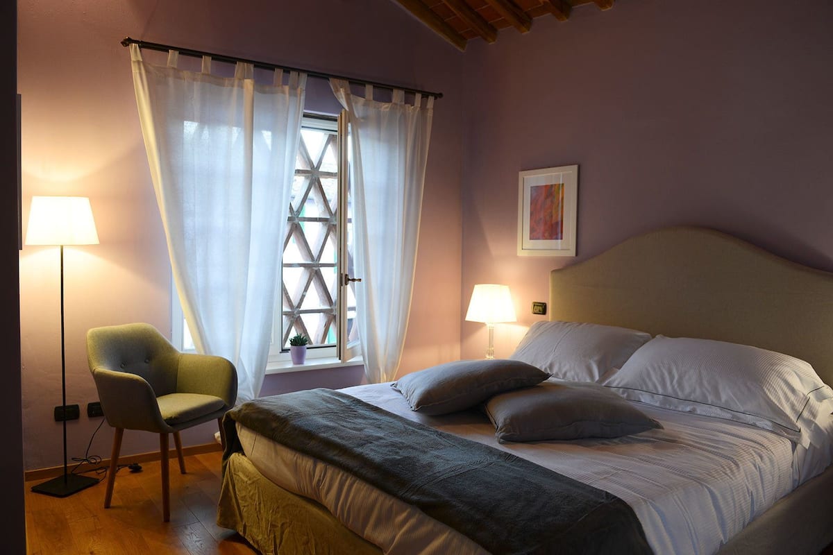 Un dormitorio con cama, una silla, ventana con cortinas, lámpara y un cuadro en la pared.