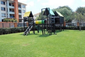 A playground with a slide and climbing structures, surrounded by a well-maintained lawn and a residential building in the background.