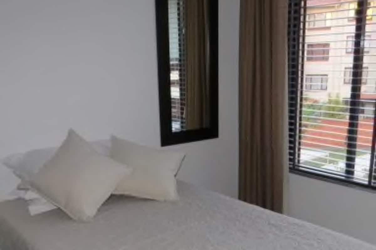 A bed with a white bedspread and two pillows, a window with blinds, and a mirror on the wall.