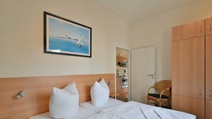 Una cama bien tendida con sábanas y almohadones blancos, un cuadro enmarcado de un velero en la pared y un armario de madera.