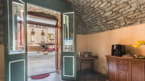 Une porte ouverte donne sur une cour intérieure dotée d’un plafond en pierre et offrant une vue sur les bâtiments environnants. À l’intérieur, on trouve un meuble en bois contenant une machine à café et une petite table.