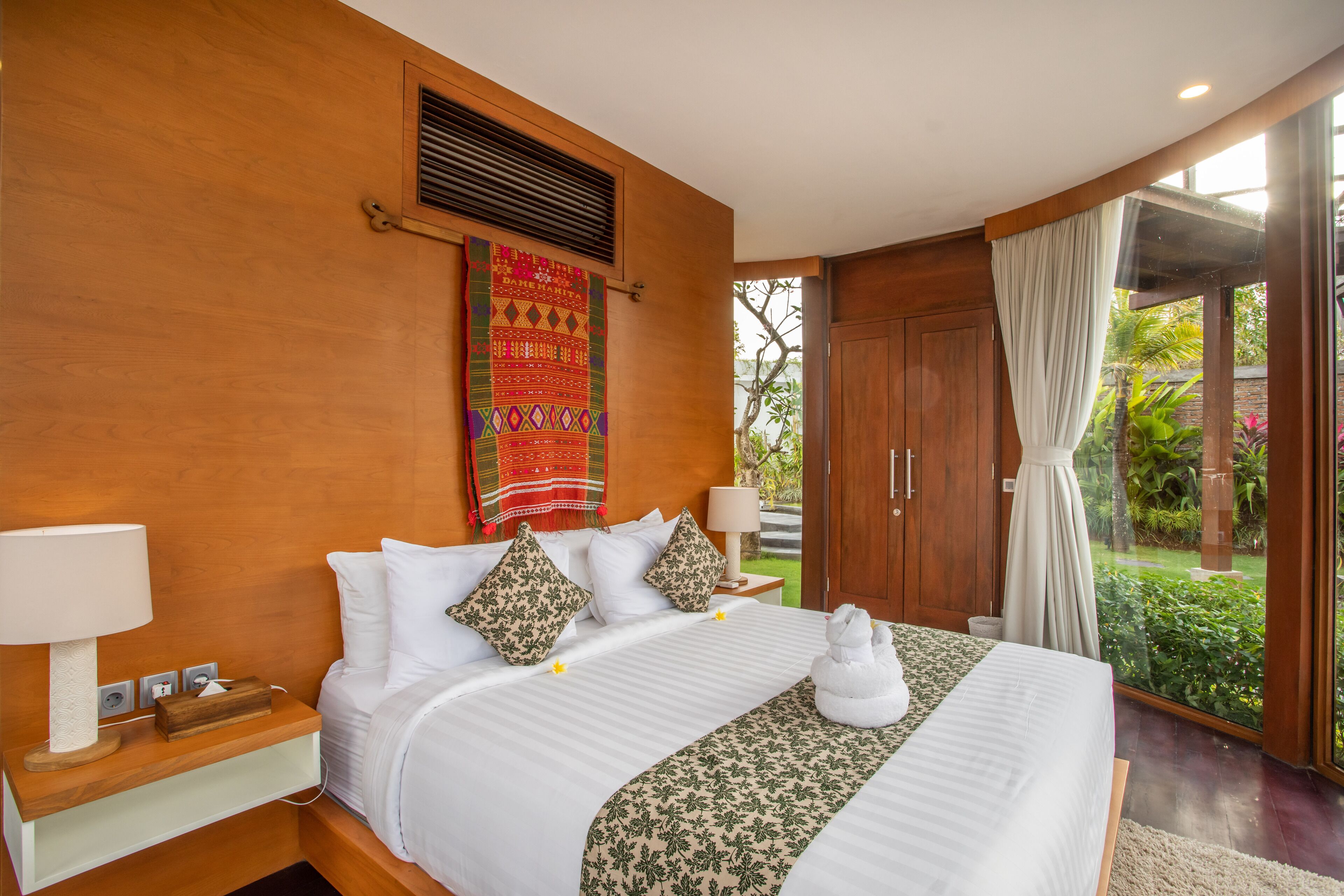 A hotel room with a wooden wall, a bed with white bedding, a decorative wall hanging, a window with curtains, and a view of greenery outside.