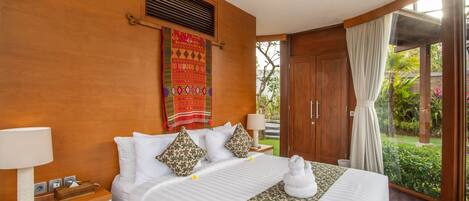 A hotel room with a wooden wall, a bed with white bedding, a decorative wall hanging, a window with curtains, and a view of greenery outside.