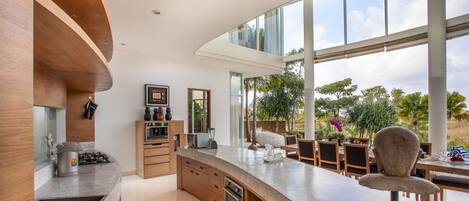 A modern kitchen with a curved island, wooden cabinets, and a large window overlooking a garden.