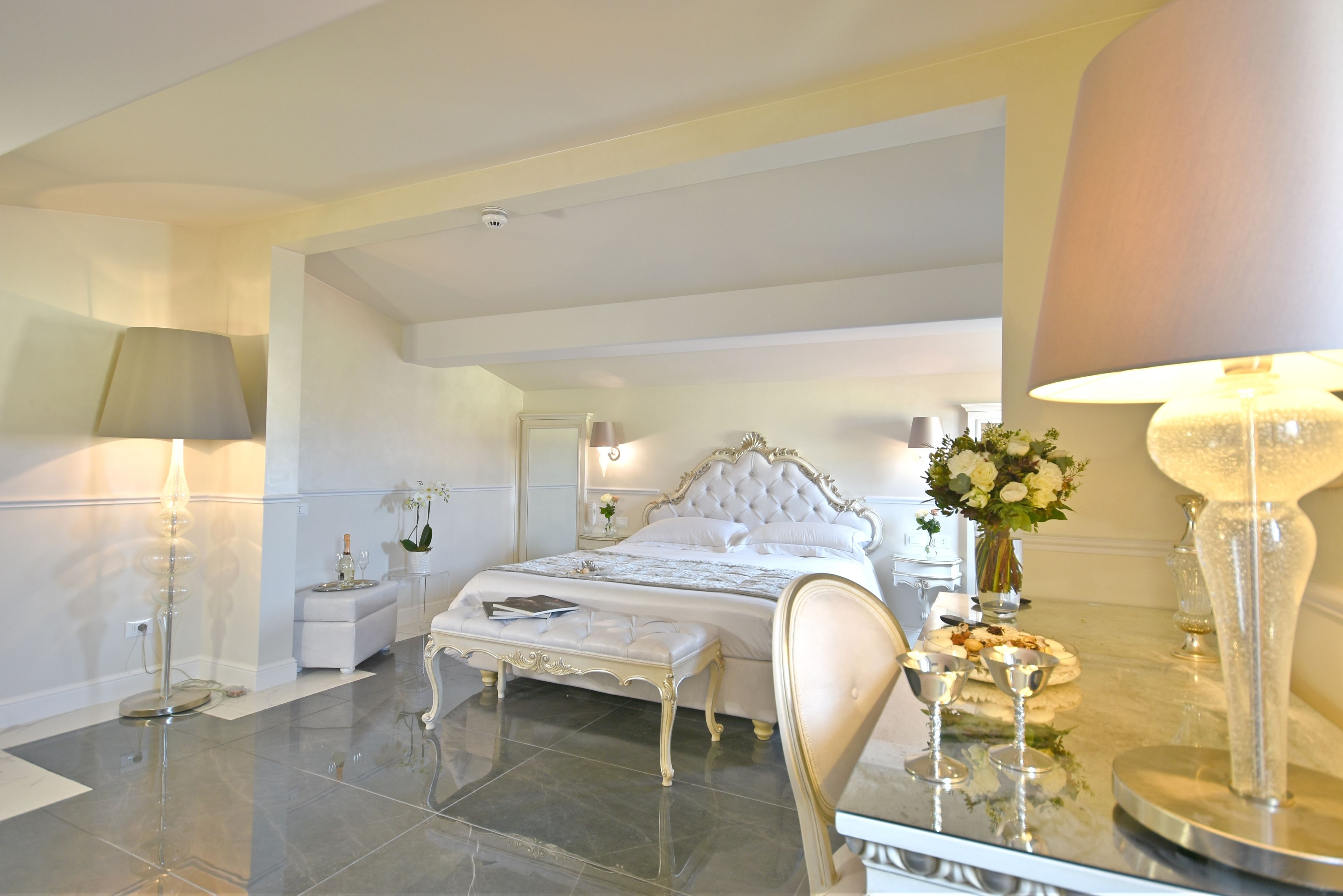 Un dormitorio lujoso con una cama grande, una mesita de noche, un espejo y una mesa de cristal con objetos decorativos.