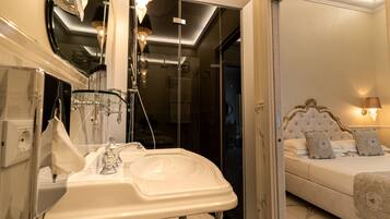 A modern bathroom with a glass shower enclosure, a white sink with a mirror, and a bedside table with a lamp.