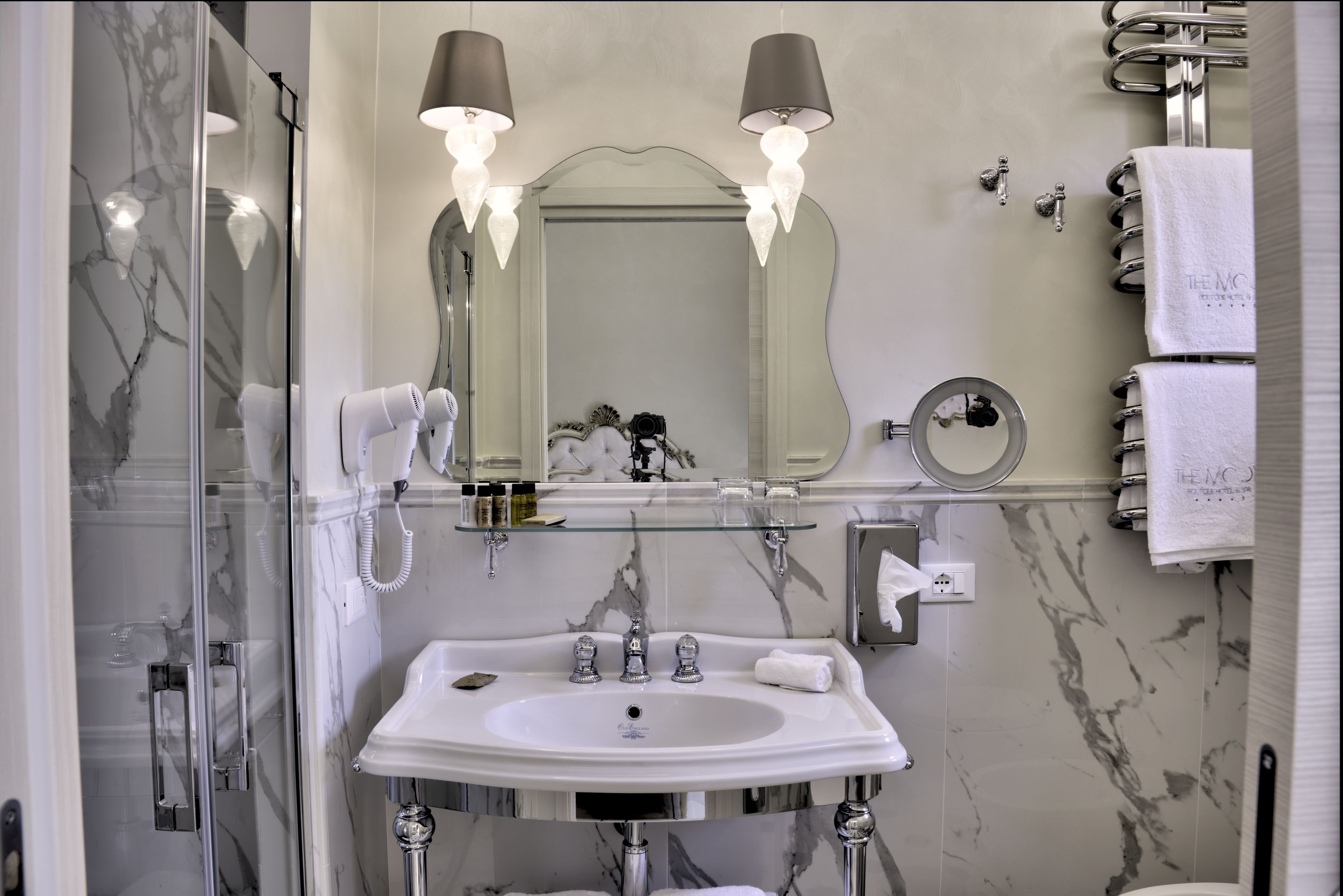 A modern bathroom with a large mirror, two wall-mounted lights, a glass-enclosed shower, a white sink with chrome fixtures, and a towel rack with white towels.