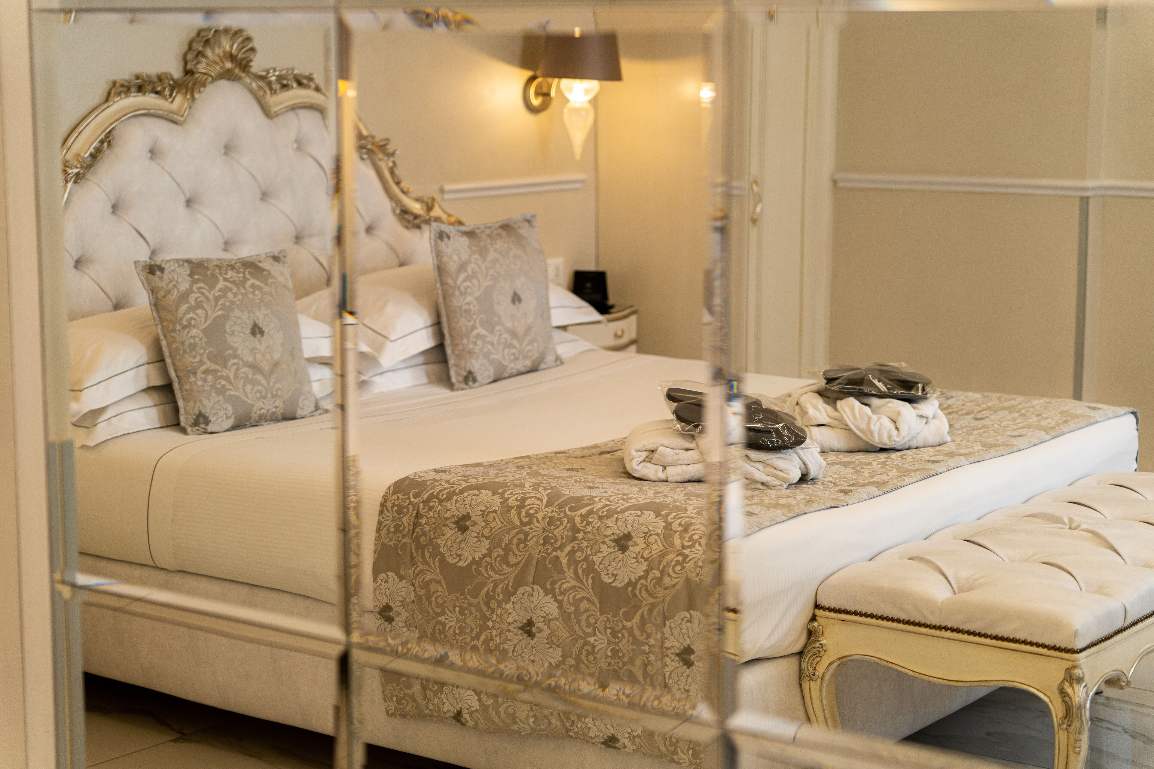 Un dormitorio lujoso con cabecera capitonada, almohadas decorativas, una cama grande con colcha estampada, una mesita de noche con lámpara y un banco al pie de la cama.