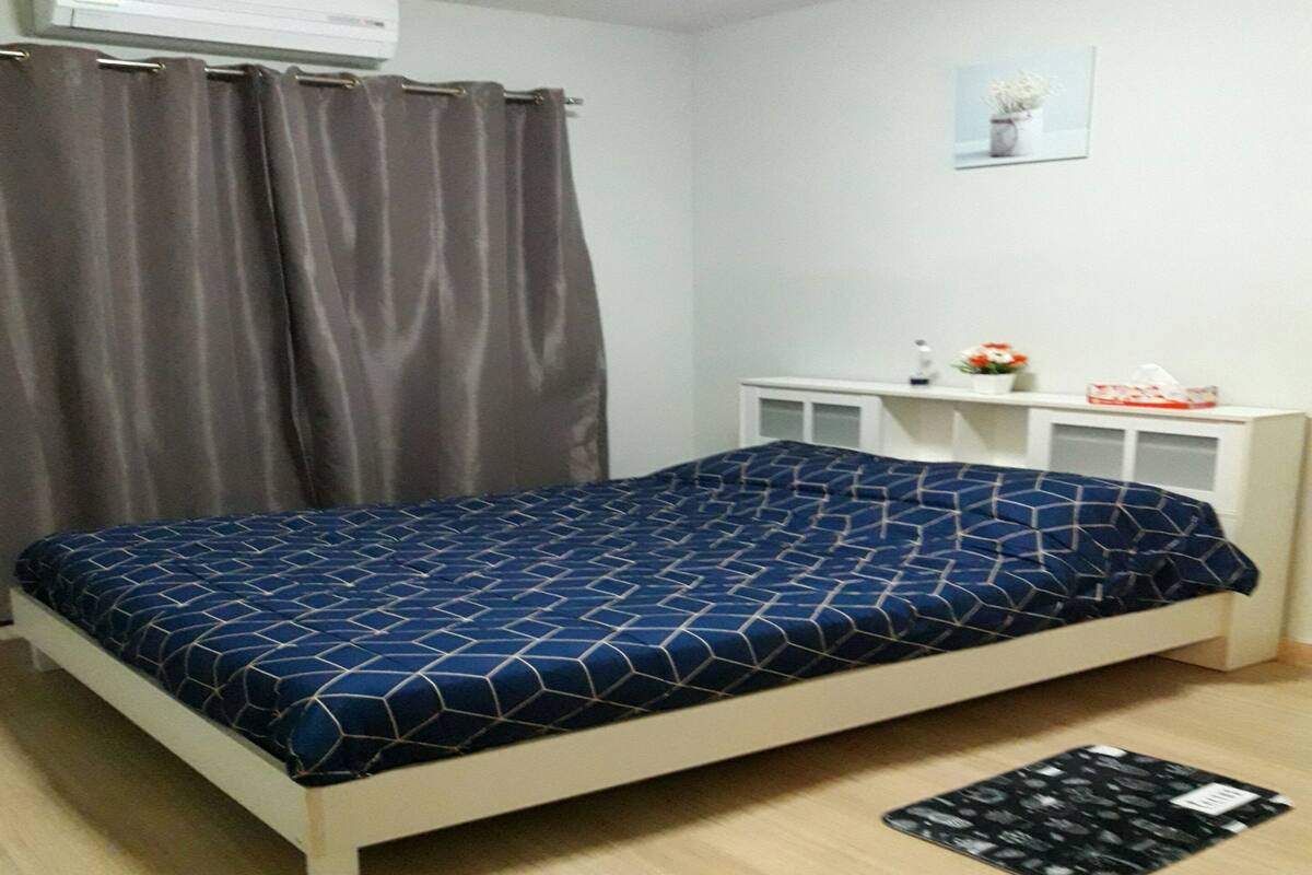 A bedroom with a bed, a white bedside table, a small white shelf, a grey curtain, and a small framed picture on the wall.