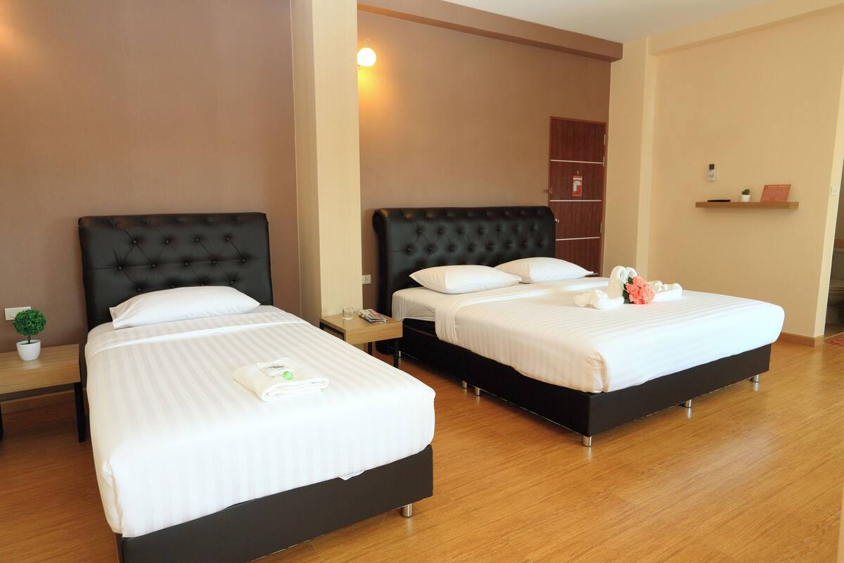 A hotel room with two beds, each with a black headboard and white bedding. There is a small table with a plant and a tray of toiletries. The room has wooden flooring and beige walls.