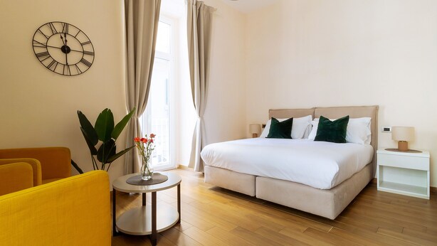 A hotel room with a bed, a round table with a vase of flowers, a yellow armchair, and a wall clock. - Musto Suites & Rooms (Naples)