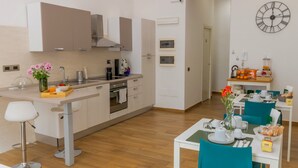 A modern kitchen with a central island, a dining area with teal chairs, and a wall clock. - Musto Suites & Rooms (Naples)