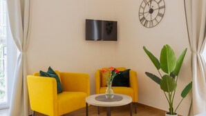 A room with two yellow armchairs, a round table with a vase of flowers, a wall-mounted clock, and a large potted plant. - Musto Suites & Rooms (Naples)
