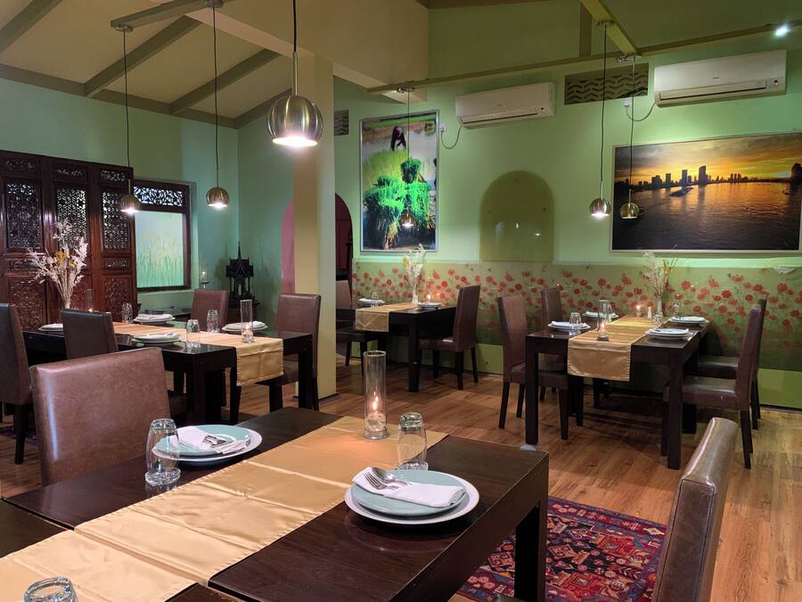 A restaurant with wooden tables and chairs, a green wall with floral wallpaper, and a cityscape painting.