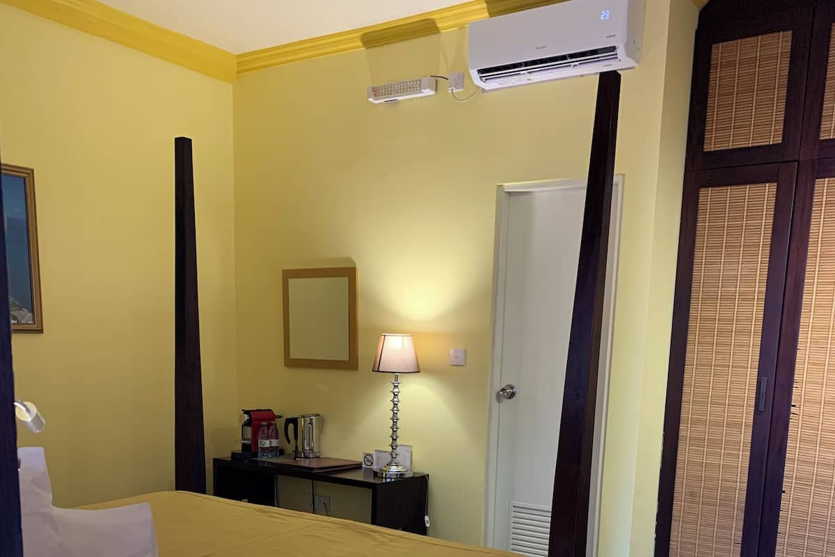 A hotel room with a bed, a bedside table with a lamp, a small table with a device, a mirror, and an air conditioning unit.
