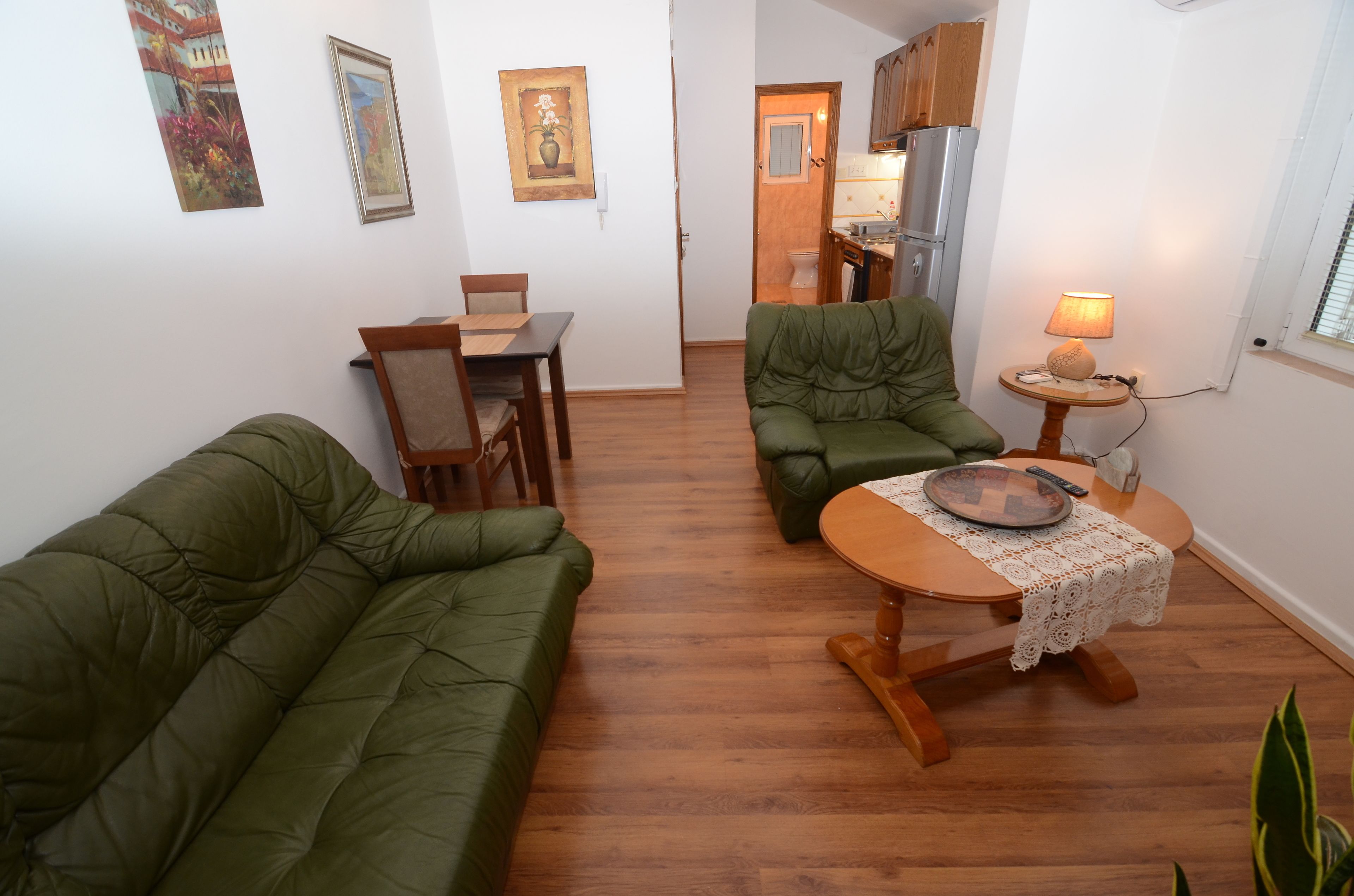 A compact living space with a green sofa, a dining table with chairs, a small round table with a lamp, and a kitchen area visible through an open door.