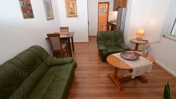 A compact living space with a green sofa, a dining table with chairs, a small round table with a lamp, and a kitchen area visible through an open door.