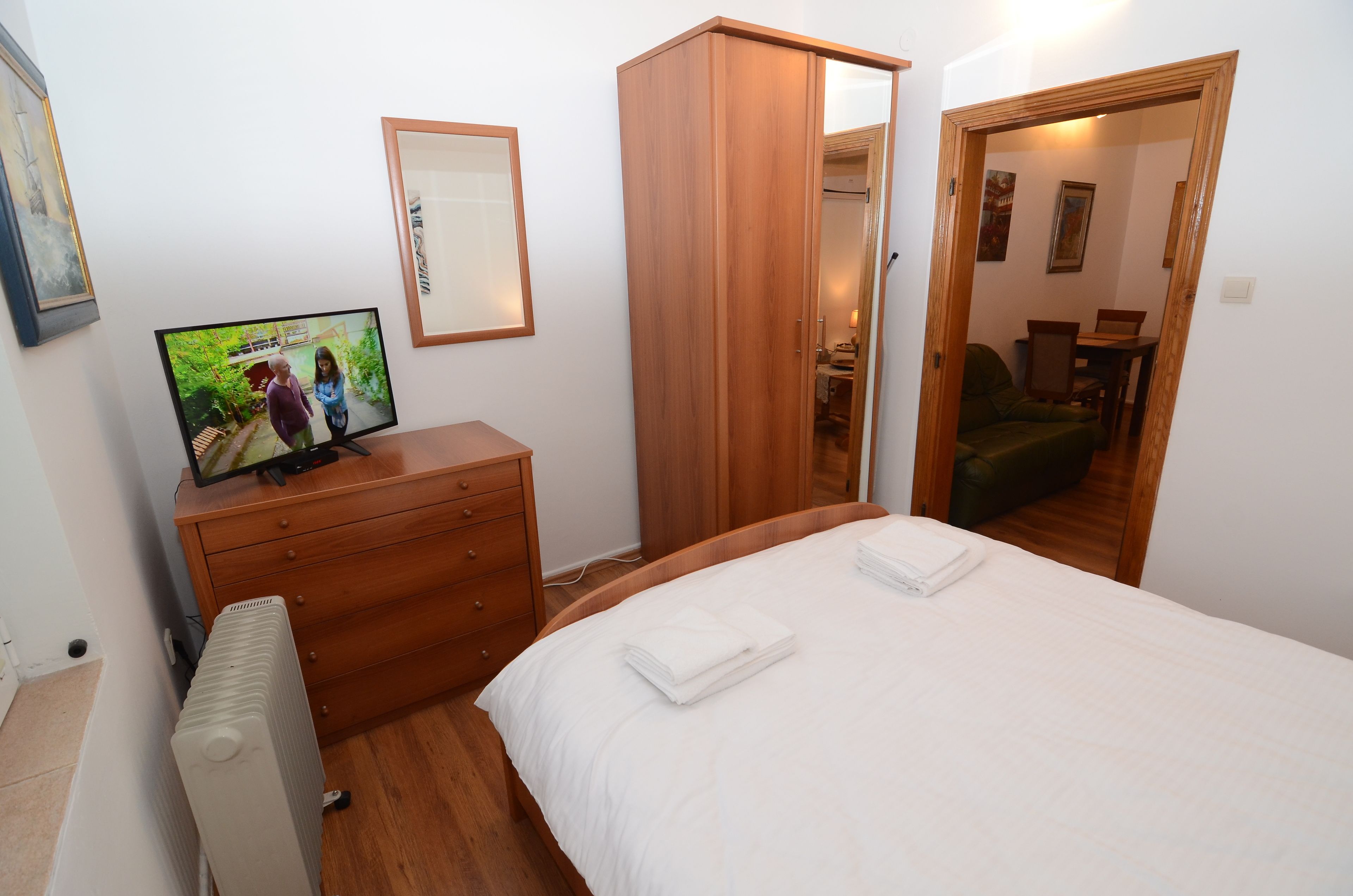 Un dormitorio con cama, armario de madera, cómoda, radiador y televisor mostrando una escena.