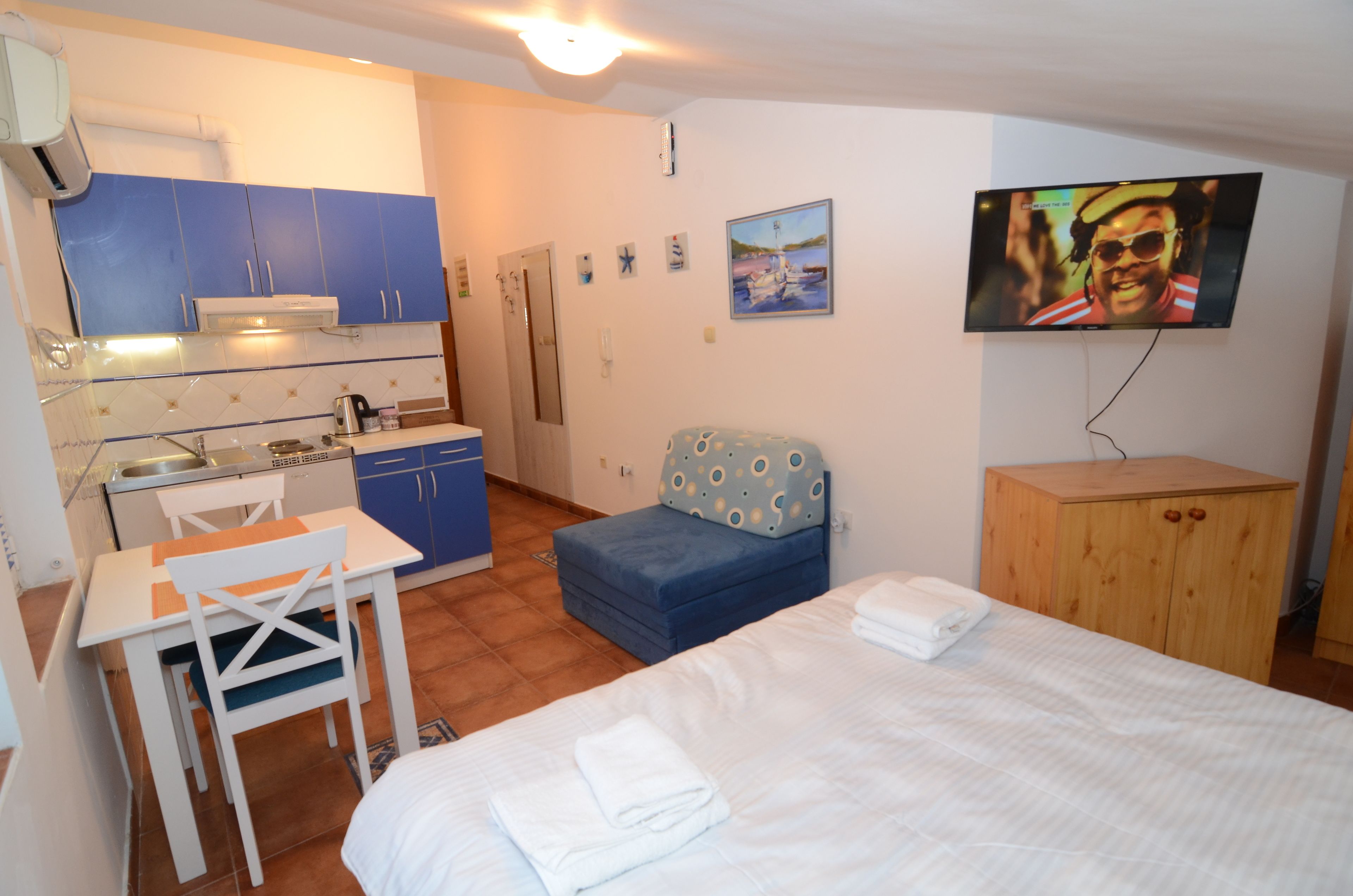 A compact hotel room with a kitchenette, a bed, a small seating area, and a television.