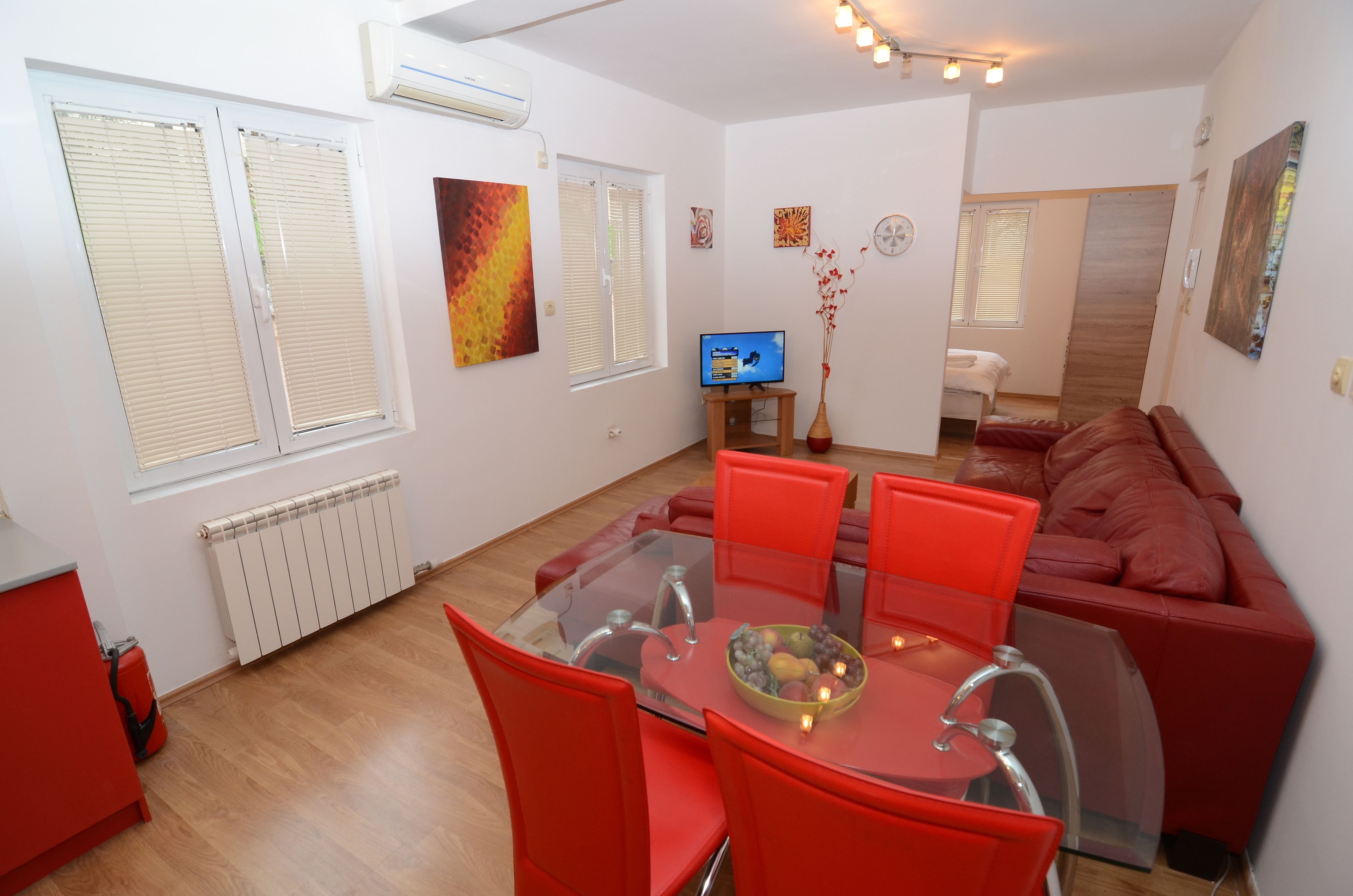 A modern living room with a glass dining table, red chairs, a red sofa, and a television.