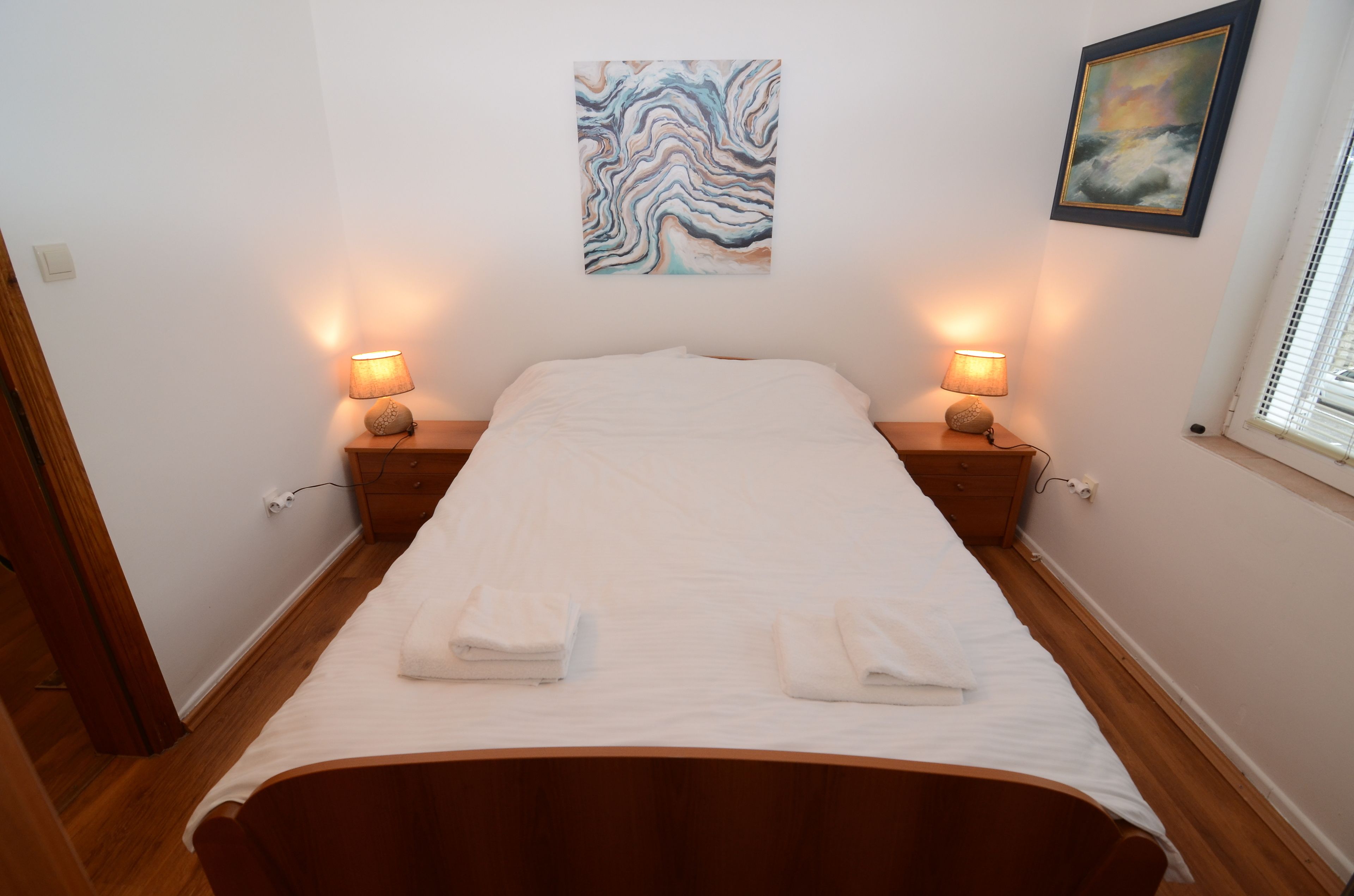 Cama bem arrumada com duas luminárias de apoio, uma obra de arte na parede e uma janela com persianas.