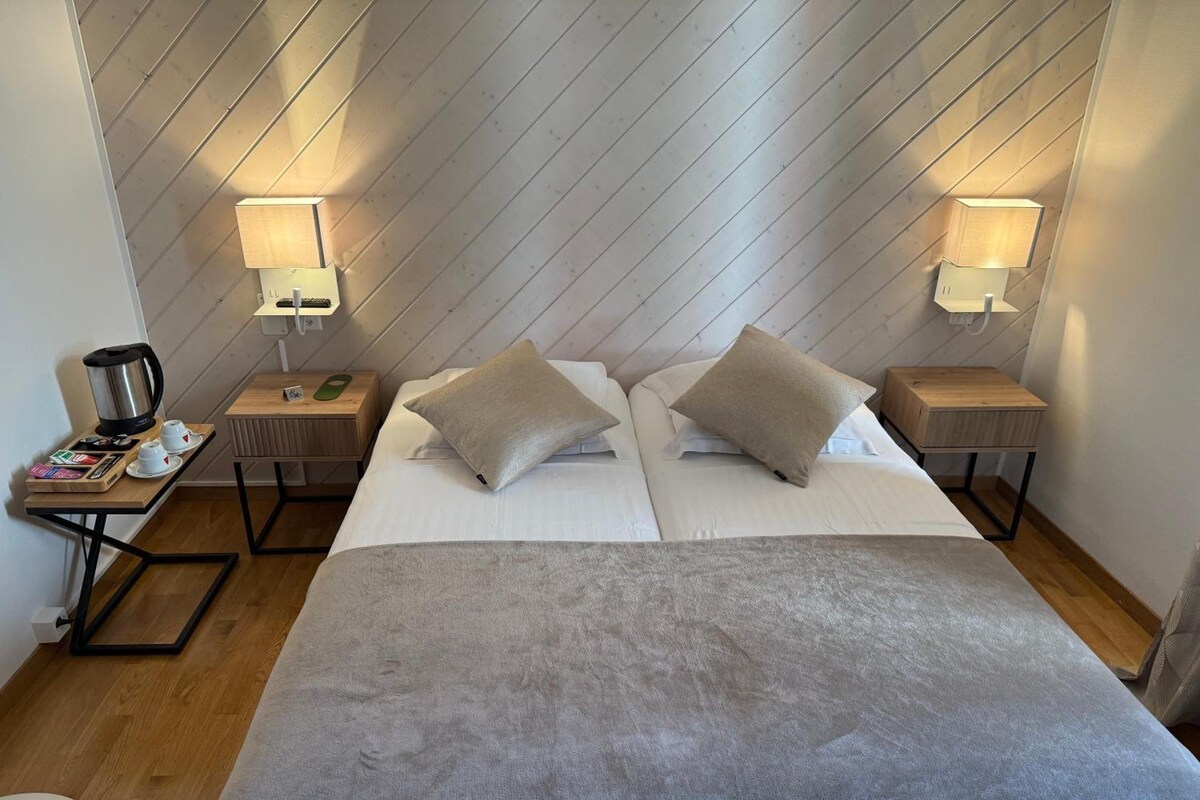 Une chambre d’hôtel avec deux lits, une table de chevet avec une lampe, une machine à café et une petite table avec des livres et une télécommande.