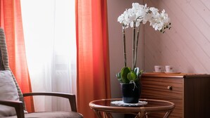 Ein Zimmer mit einem Sessel, einem kleinen runden Tisch mit einem Blumentopf, einem Holzschrank und einem Fenster mit Vorhängen.