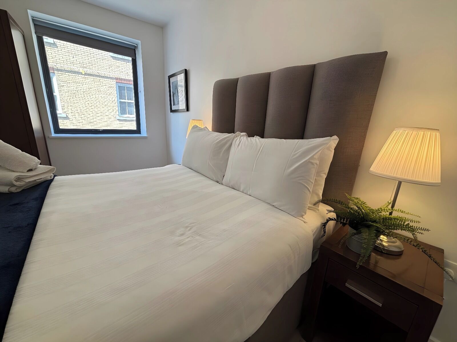 A neatly made bed with white linens, a bedside lamp, and a view of a brick building through a window.