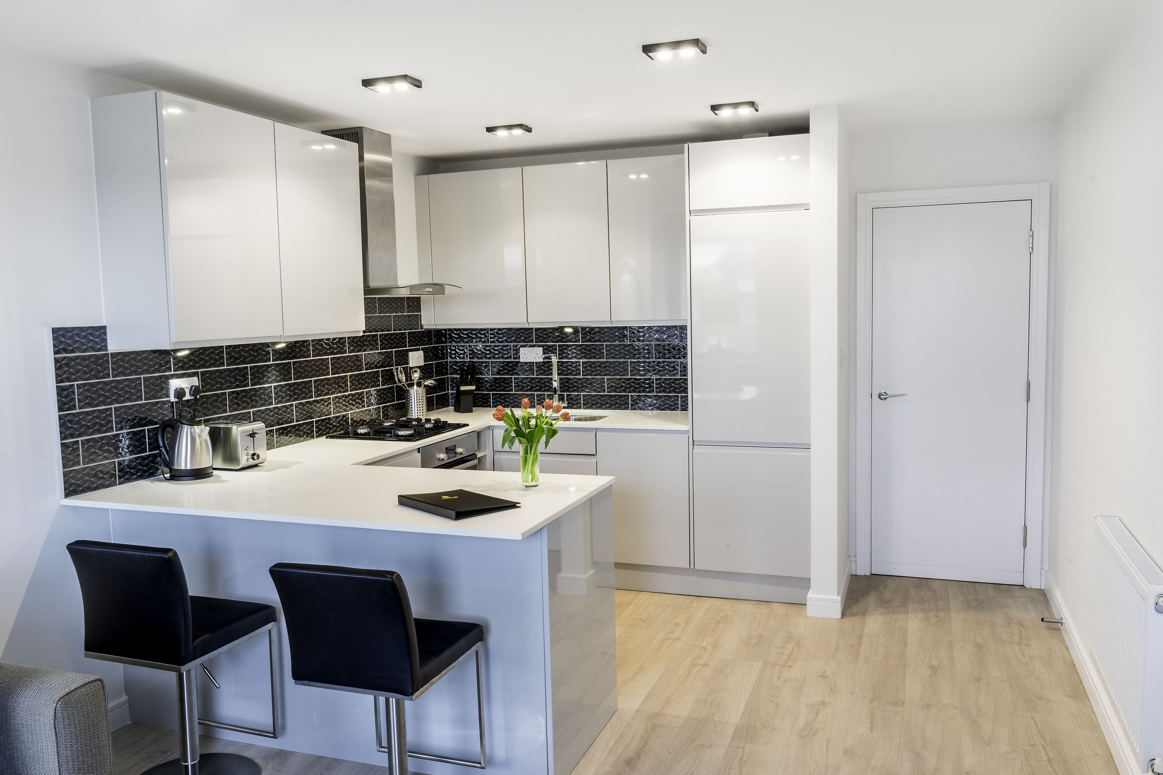 A modern kitchen with white cabinets, a black and white backsplash, a central island with bar stools, and a door leading to another room.