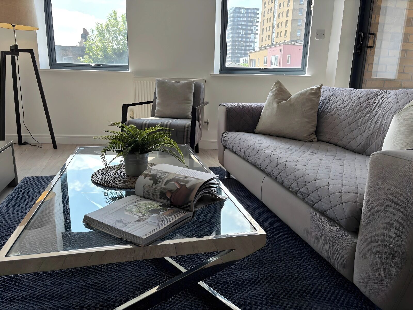 A modern living room with a glass coffee table, a grey sofa, and a view of buildings through large windows.