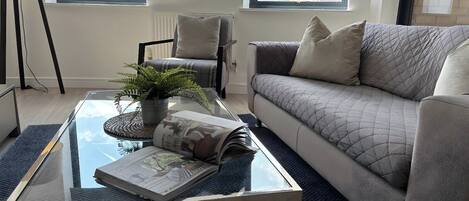 A modern living room with a glass coffee table, a grey sofa, and a view of buildings through large windows.