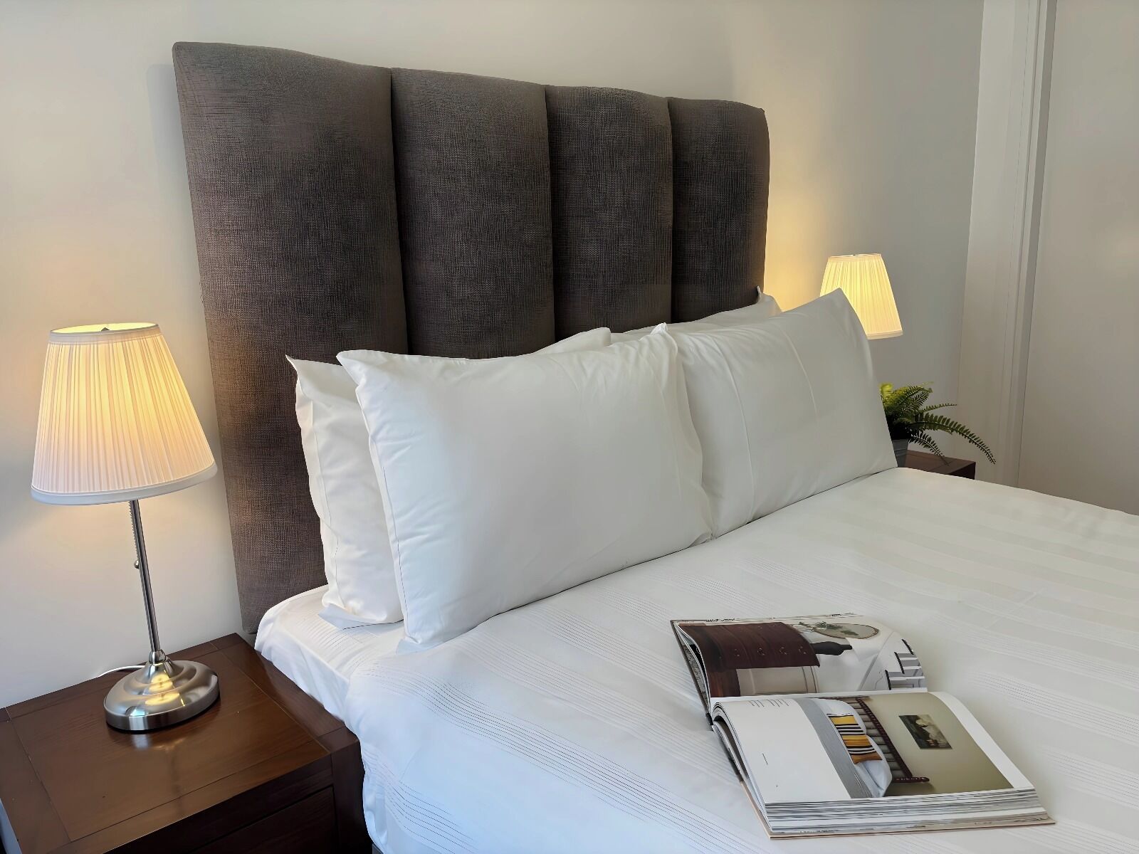 A neatly made bed with white linens and pillows, a bedside lamp, and a magazine on the nightstand.