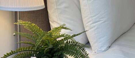 A neatly made bed with white linens, a bedside lamp, a potted fern, and a stack of books on a nightstand.