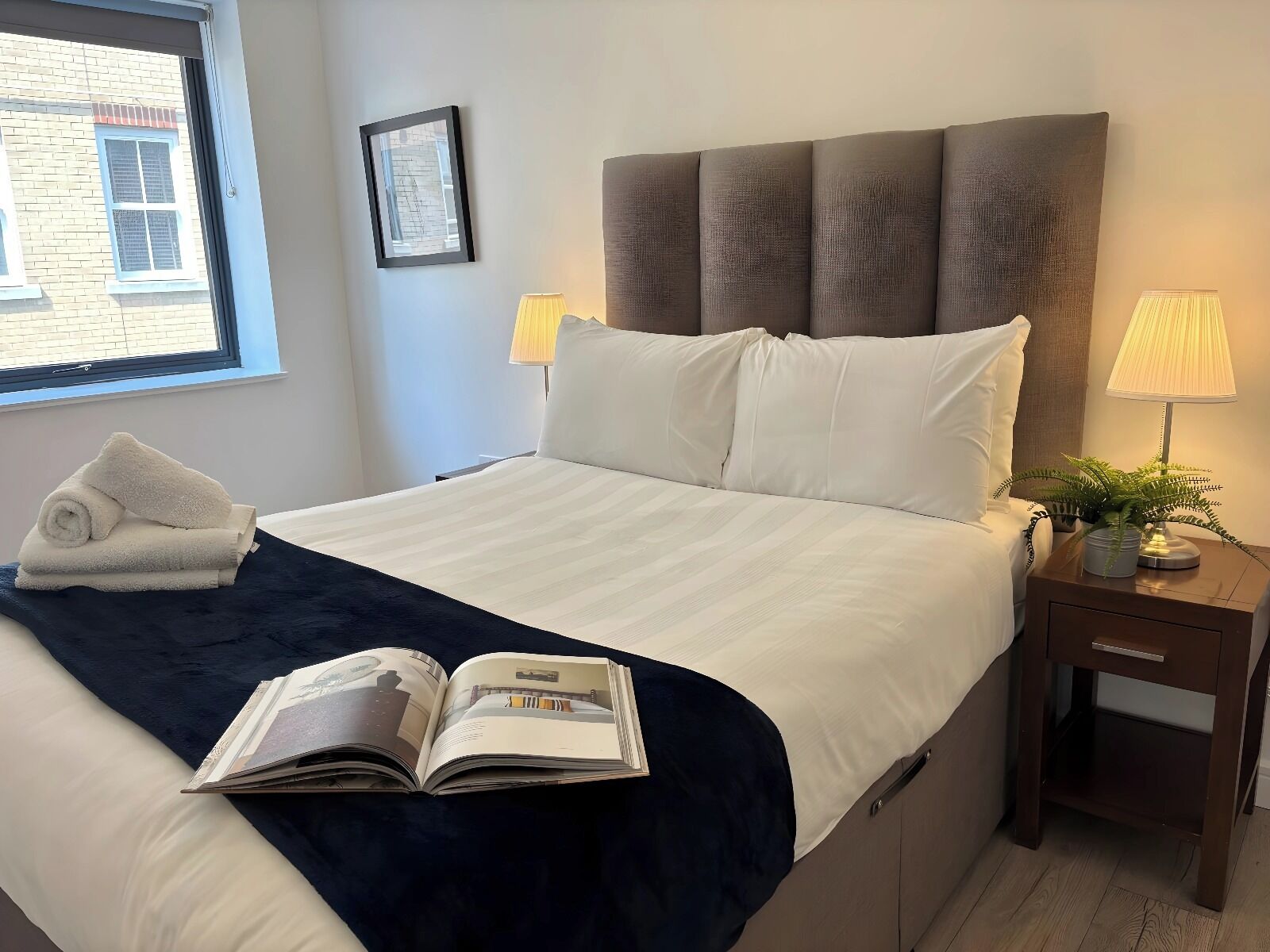 A neatly made bed with a dark blue throw blanket, a rolled white towel, and an open magazine on the bed. There is a nightstand with a lamp and a potted plant beside the bed.