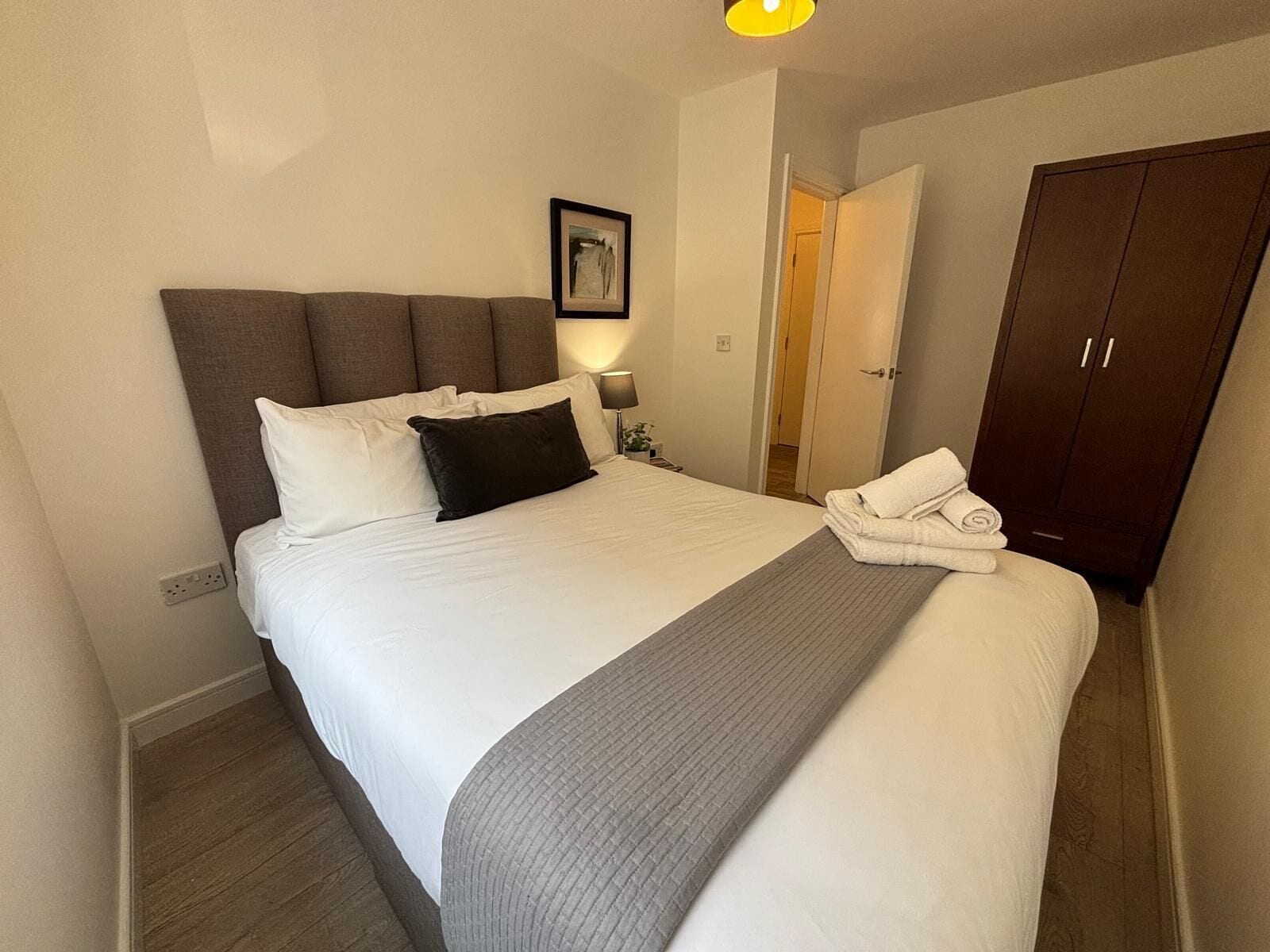 Una cama bien hecha con almohadas blancas y oscuras, una manta gris y una toalla doblada al lado.