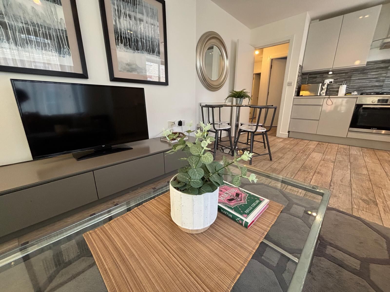 Uma sala de estar moderna com televisão de tela plana, uma mesa de centro de vidro com uma planta e um livro, uma área de refeições com cadeiras, e uma cozinha com armários e eletrodomésticos.