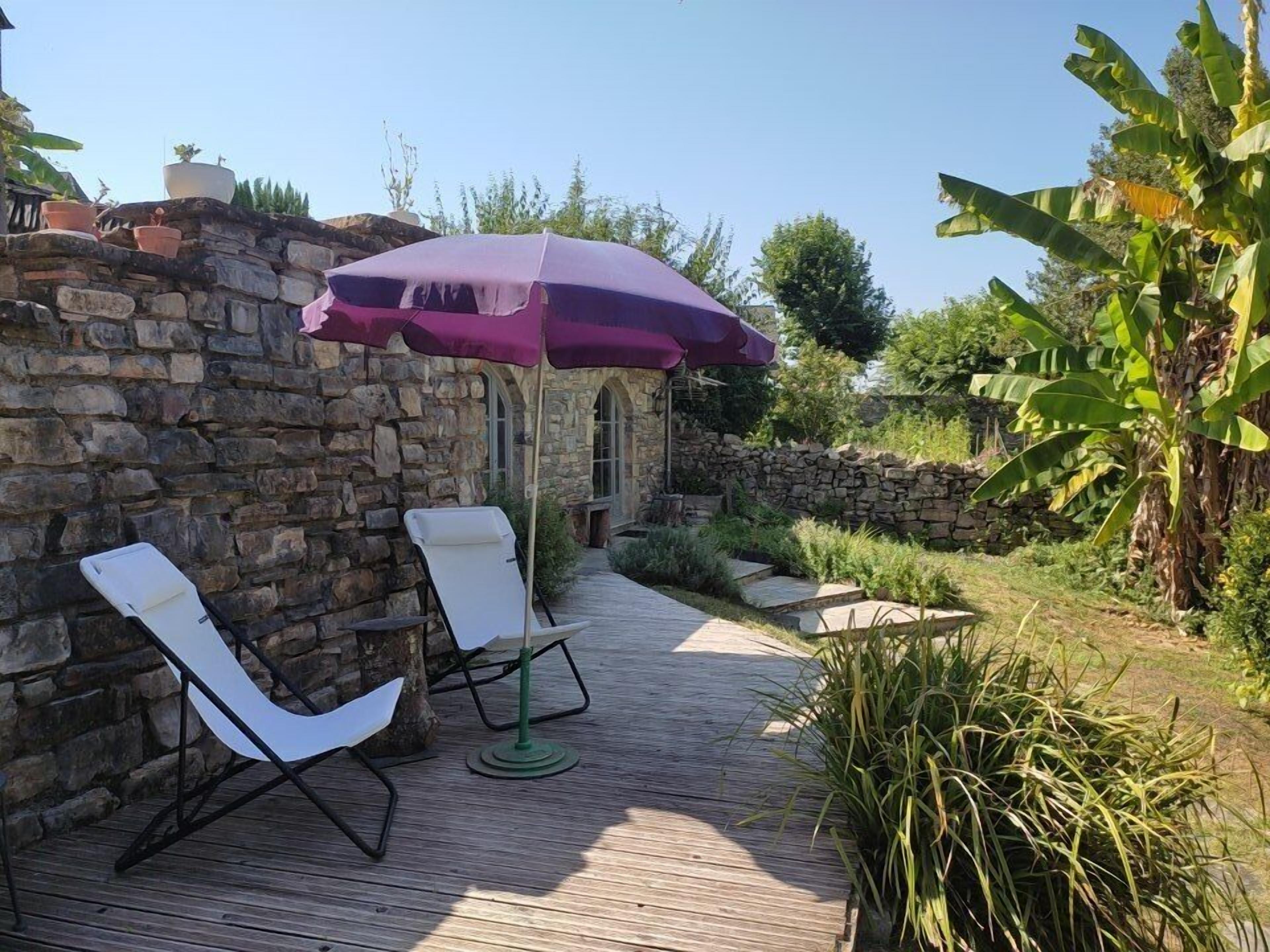 An outdoor patio with stone walls, two white lounge chairs, a purple umbrella, and a banana tree.