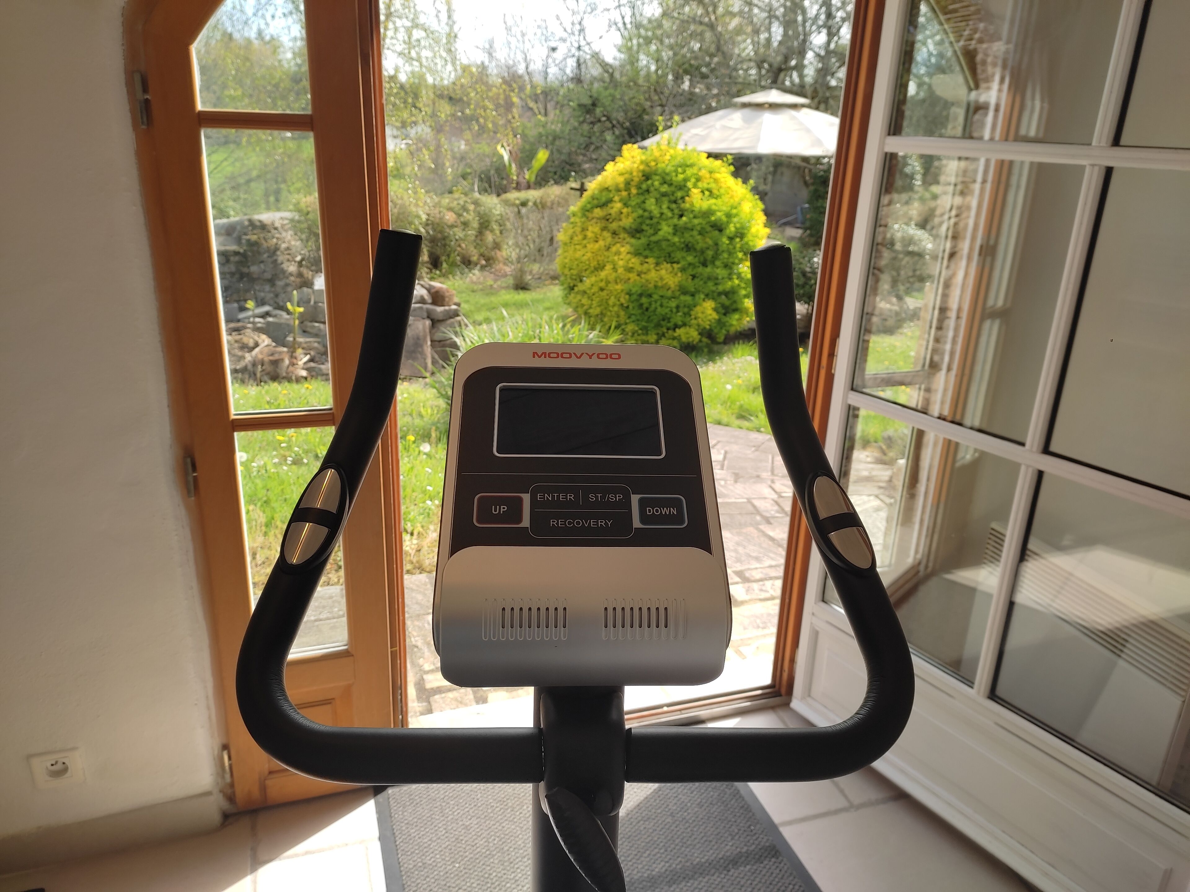 Una bicicleta estática con pantalla digital, ubicada cerca de una ventana con vistas a un jardín.