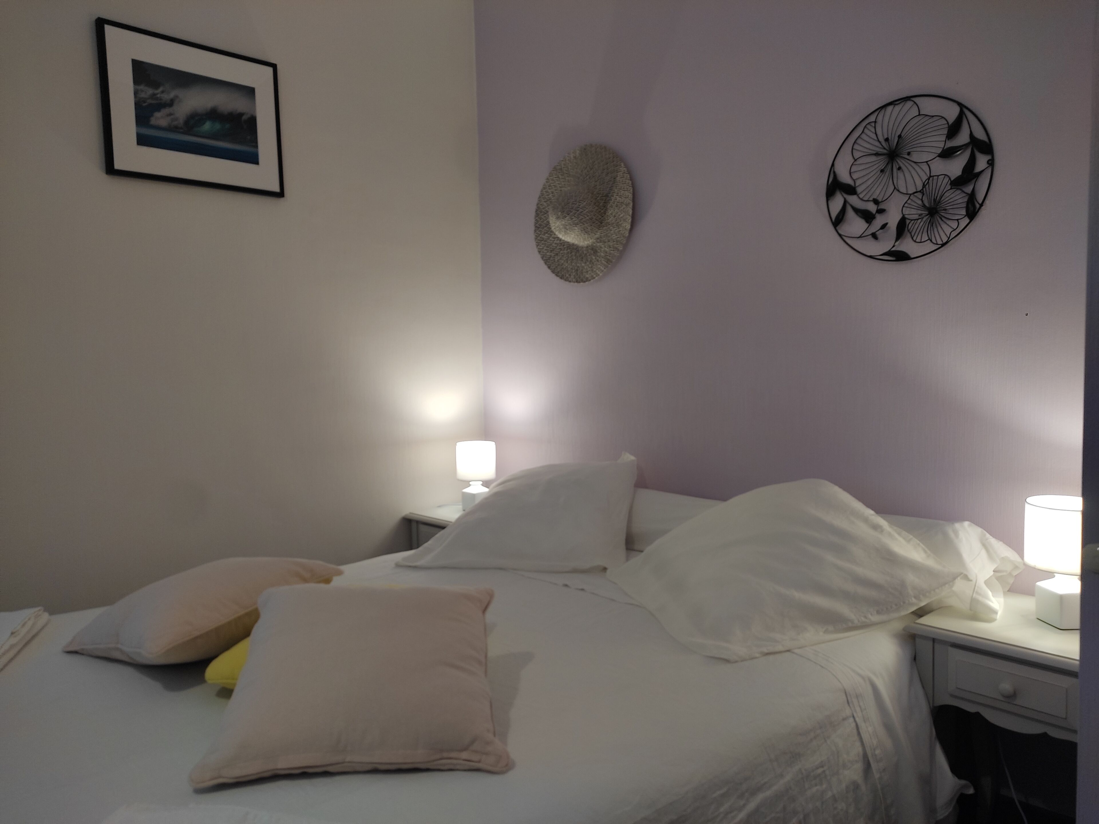 Een net opgemaakt bed met witte lakens en meerdere kussens, een nachtkastje met een lamp, en een decoratief object aan de muur.