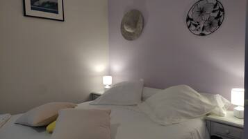 Een net opgemaakt bed met witte lakens en meerdere kussens, een nachtkastje met een lamp, en een decoratief object aan de muur.
