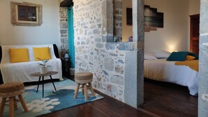 A cozy room with a stone wall, a wooden floor, a white sofa with yellow pillows, a small wooden stool, and a bed with a blue pillow.