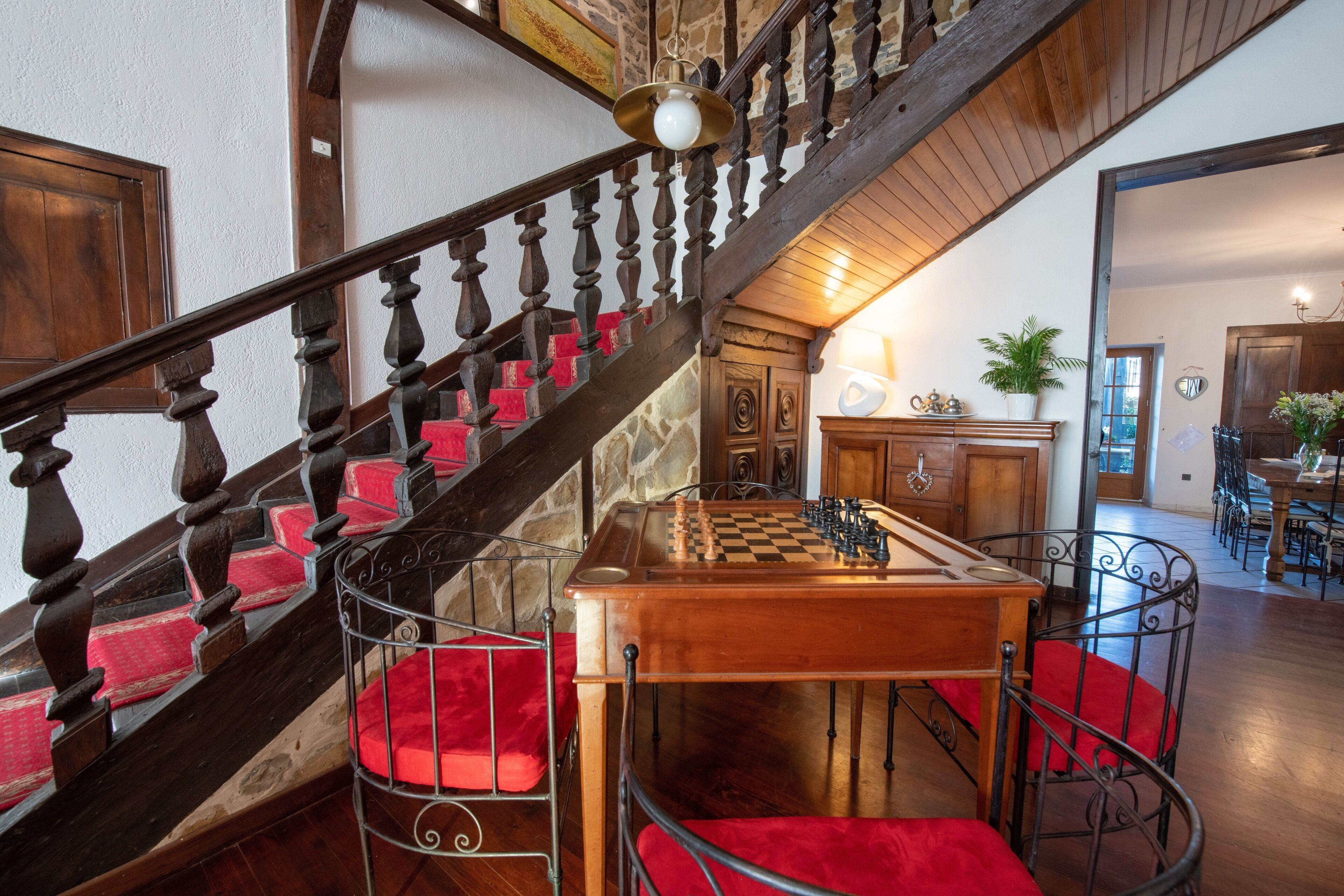 Un escalier en bois avec des marches recouvertes d’un coussin rouge, une table en bois avec un jeu d’échecs, et un coin repas avec des chaises et une table.