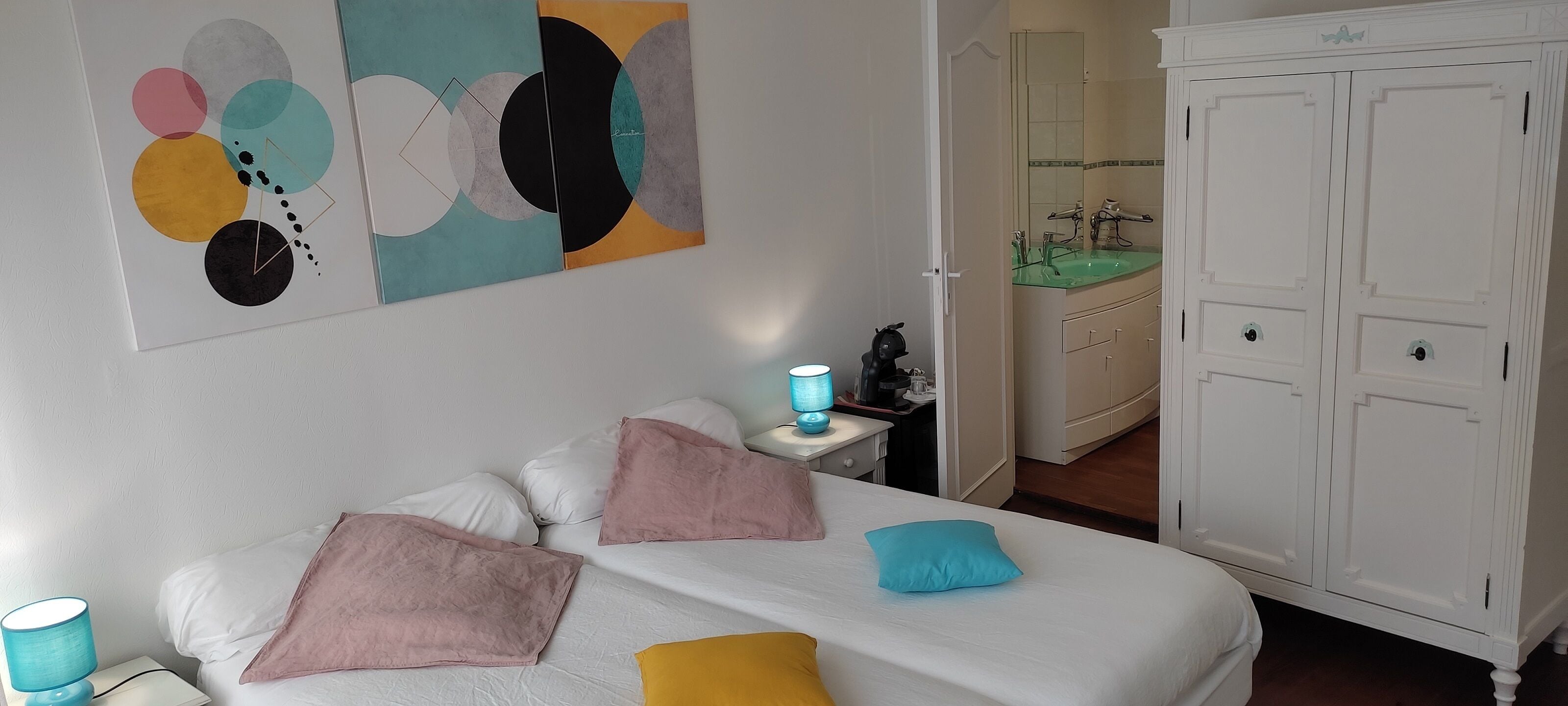 A bedroom with a bed, pillows, a nightstand, a lamp, a wardrobe, and a bathroom visible in the background.
