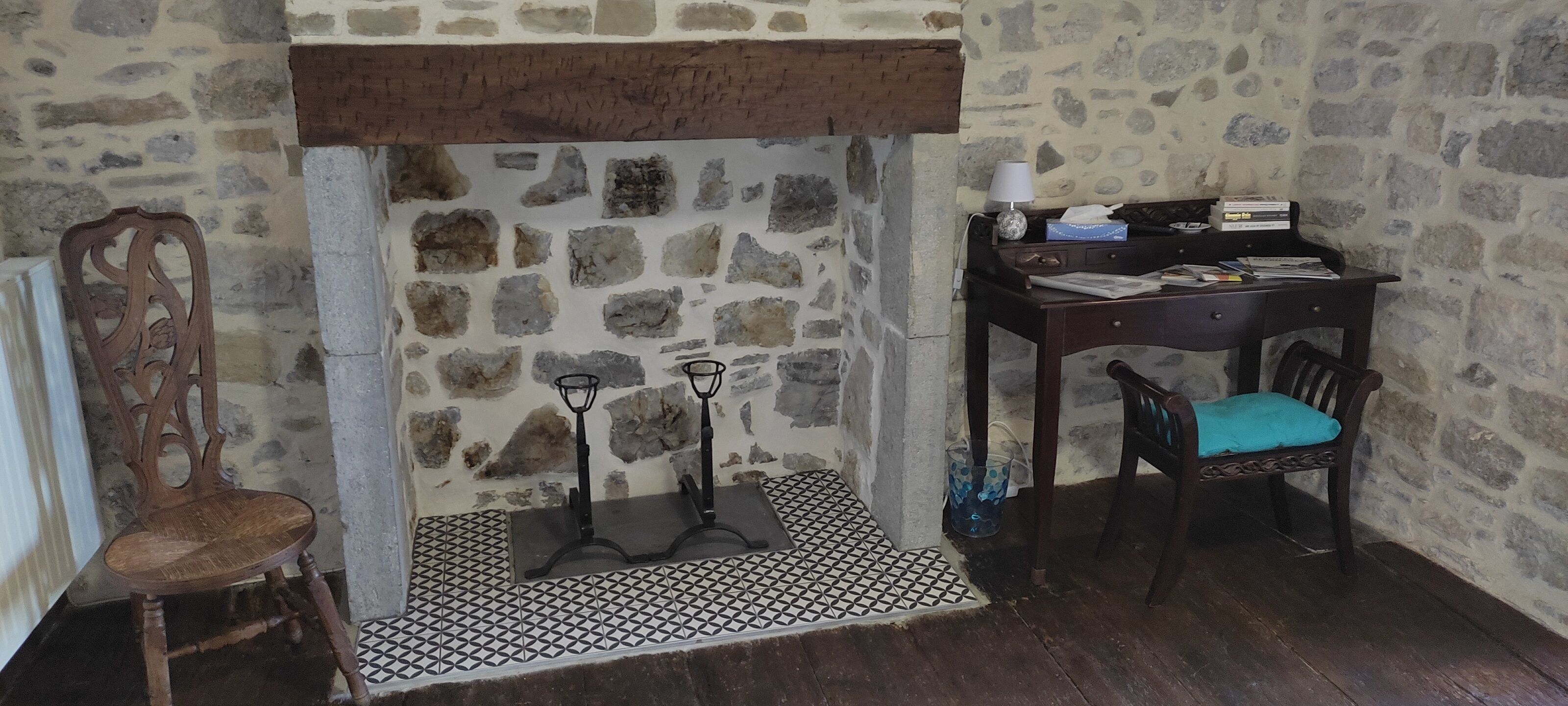 Habitación con pared de piedra, chimenea, una silla de madera, un escritorio con lámpara y una alfombra estampada.